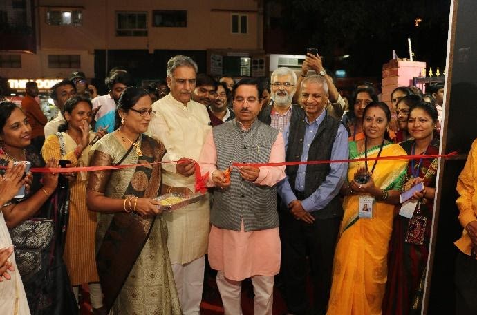 Indian man in pink long sleeves and gray vest cuts red ribbon in event with group of people around